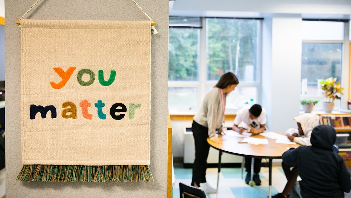 Sign that says "You Matter" hangs in a room. Teacher and students are at a table in the background.
