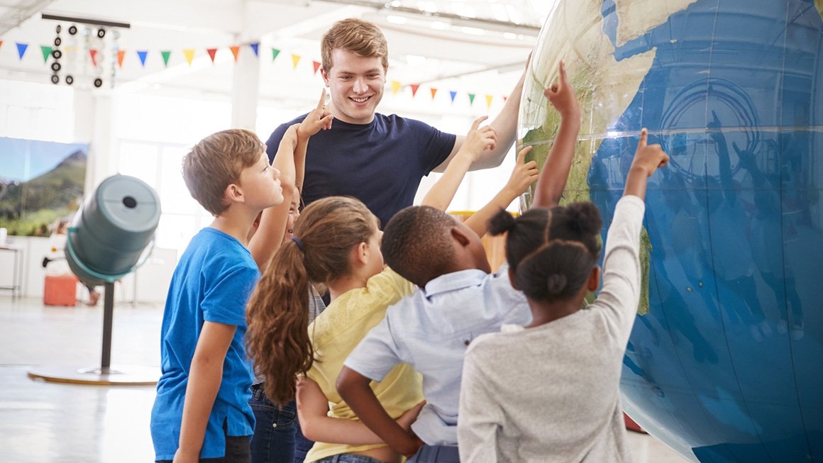 Teacher and students stand next to a huge globe and point to different parts of it. 