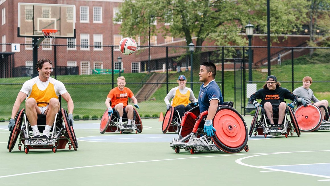Smiling students in sport wheelchairs play wheelchair rugby on a green tennis court