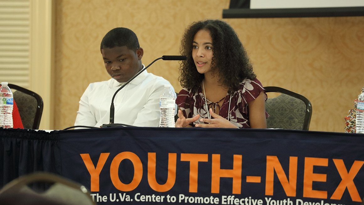 Two youth, seated behind a table with microphones on a stage, speak at the 2022 Youth-Nex conference.