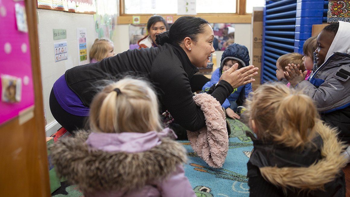 Preschool teacher sits on the floor with her students, claps her hands.