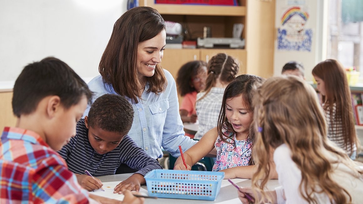 A teacher sits at a table where elementary school children work, sitting next to her.