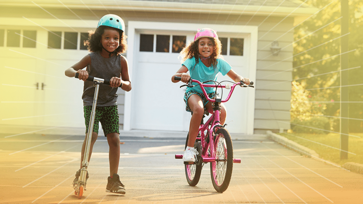 A child stands on a scooter wearing a helmet smiling next to another child on a bicycle wearing a helmet.
