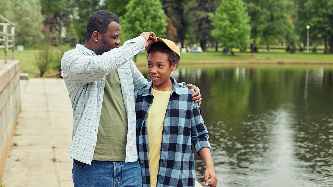 Man and boy walk on dock alongside water. Man playfully tugs on the bill of the hat the boy is wearing.