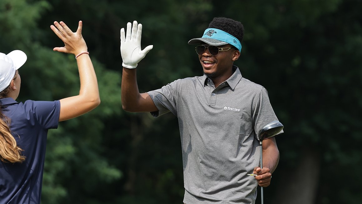 Teen wearing a visor and shirt with "First Tee" logo carrys a golf club and raises hand to give someone a high five.