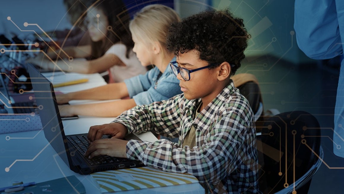 Boy student wearing glasses sits at a table in school working on a laptop.