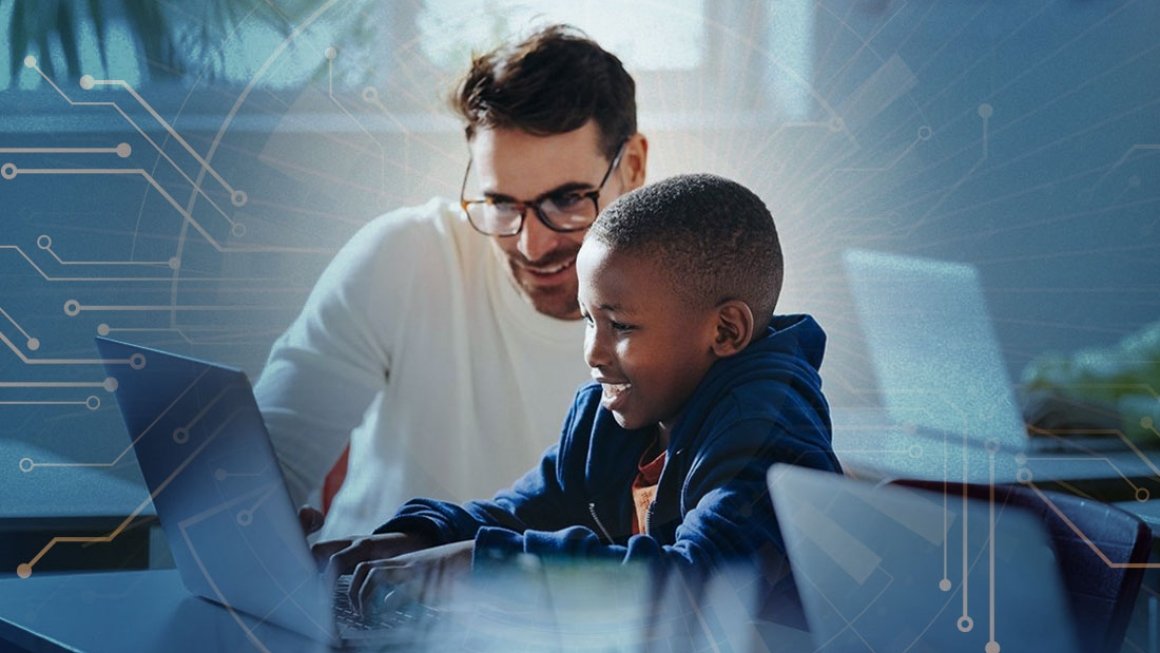 Male student sits next to young male student who works on a laptop at a desk.