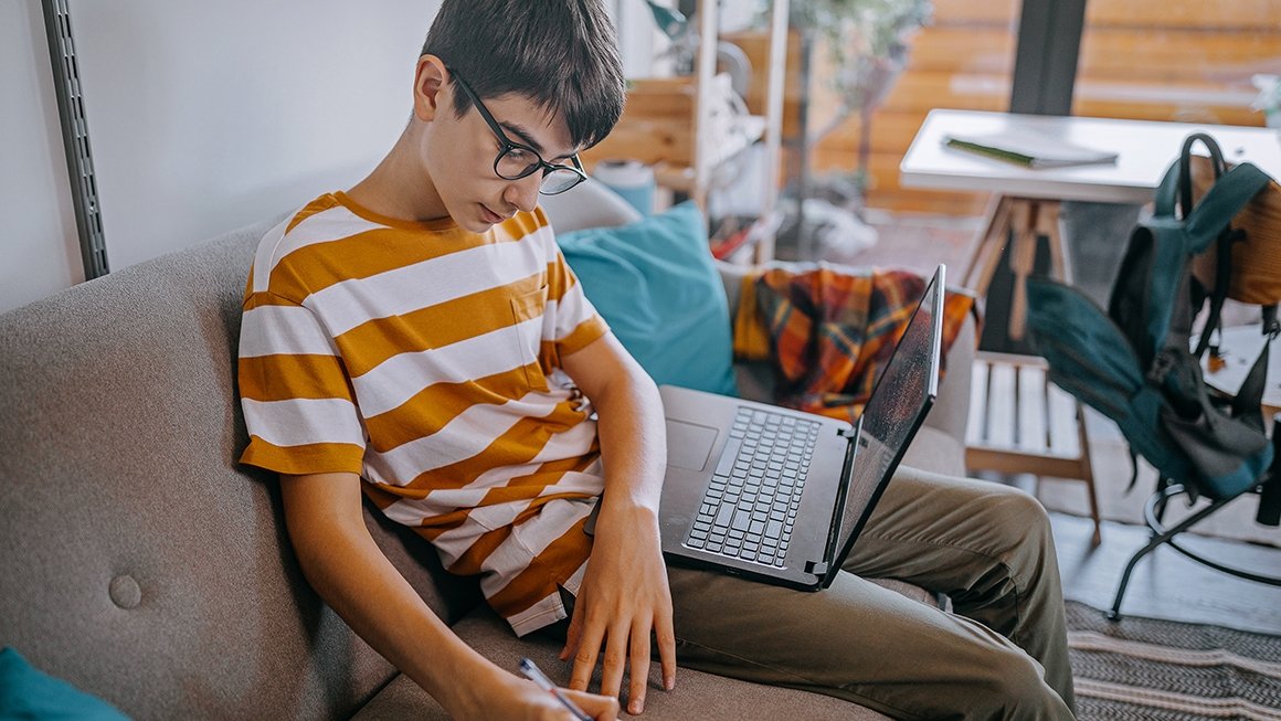 Teen boys works on homework while sitting on a couch at home.