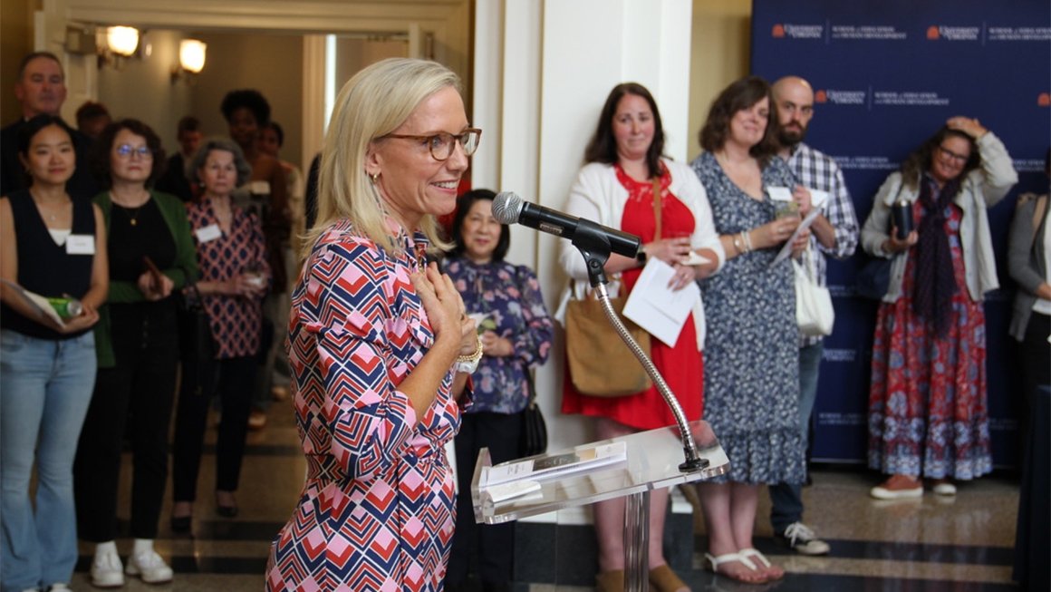 A woman with blonde hair, glasses, and a patterned dress stands at a podium smiling and placing her hand on her heart.