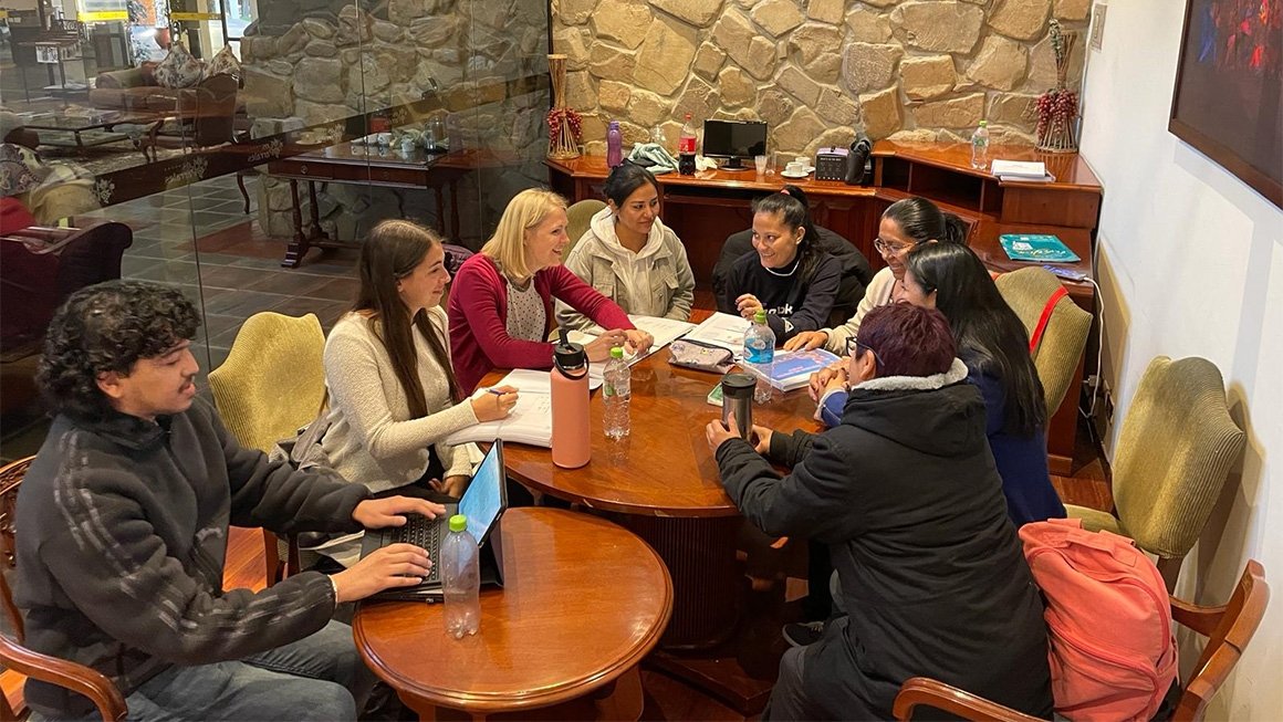 Eight people sit around round tables with notebooks and laptops