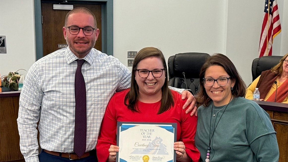 A young woman wearing glasses and a red top smiles while holding up a certificate that says "Teacher of the Year." A man and a woman stand on either side of her smiling at the camera.