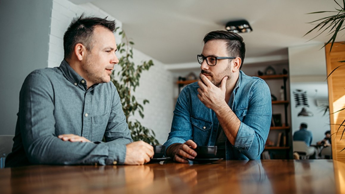 Two men sit at a table in a living room talking.