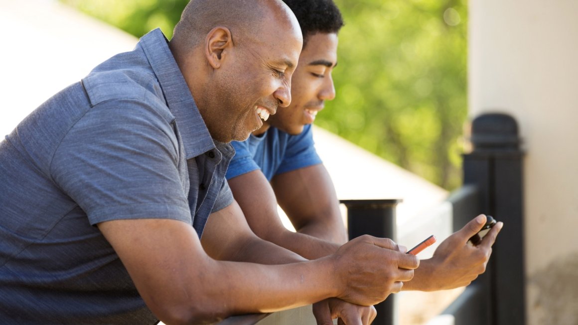 Older male leans on railing next to younger male, both smiling and looking at their phones.