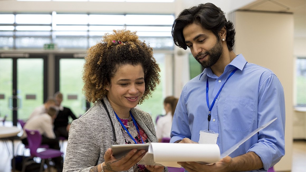 Two professionals stand in a school looking at a clipboard together