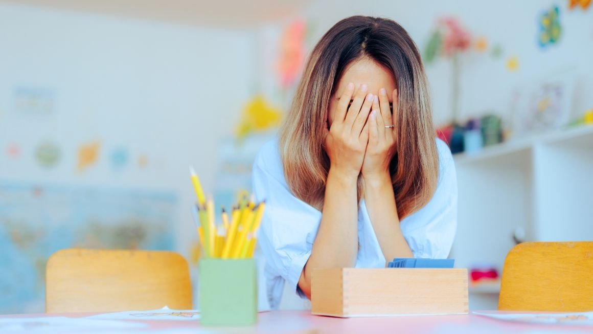 A teacher sits at a desk in a classroom with her face in her hands