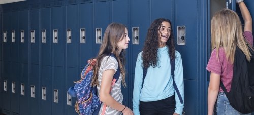 Three adolescent girls stand next to a bank of blue lockers, talking and laughing