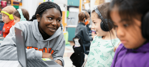 A teacher smiling at a child who is working at a laptop with headphones on
