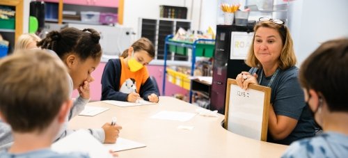 A teacher sits at a table with several elementary school students, holding up a small whiteboard that reads "twelve times five equals"