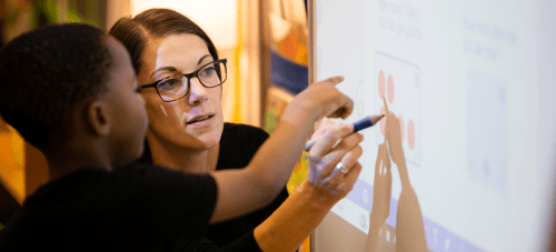 A teacher working in a classroom with a young child at a whiteboard.