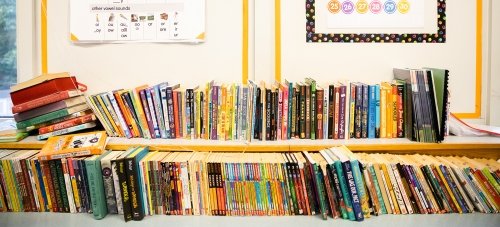 Two long rows of colorful books on a shelf in an elementary school classroom. A poster on the wall above illustrates vowel sounds.