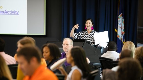 View of a larger conference room from the back of the room. Many people seated in front of view at tables. The presenter is in the back of the room.