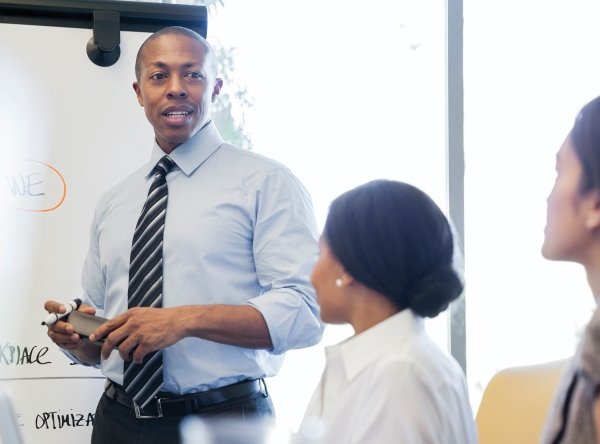 topic-checkerboard_policy-leadership_ple_2022-10-31.jpg Man in a blue shirt and tie stands at a whiteboard, facing two women who are seated at a table