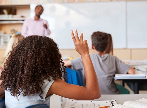 educating-for-democracy_checkerboard_2022-11-11.jpg Student in classroom raises her hand while teacher stands at front of class
