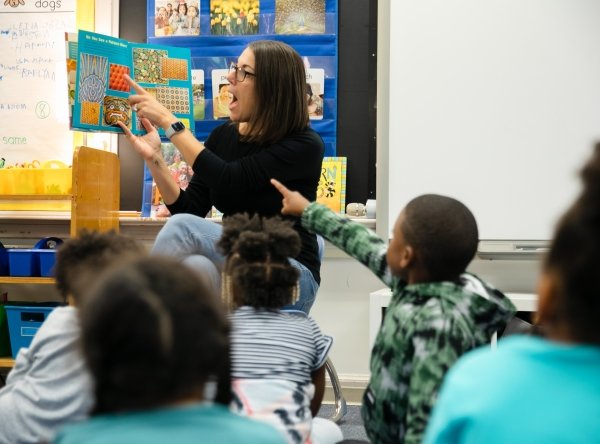 A kindergarten teacher points at pictures in a book while students, seated on a rug in front of her, look and point