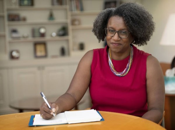 Dean Stephanie Rowley sitting at her desk writing in a notebook and looking at the camera and smiling