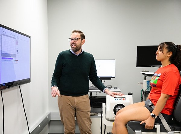 easil-lab_checkerboard_thigh-muscle-strength_2023-09-21.jpg Woman sits in chair while professor stands next to them. They both look at a screen in front of them with a graph