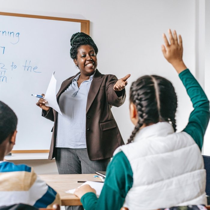 A teacher at the front of a classroom calling on a student raising their hand