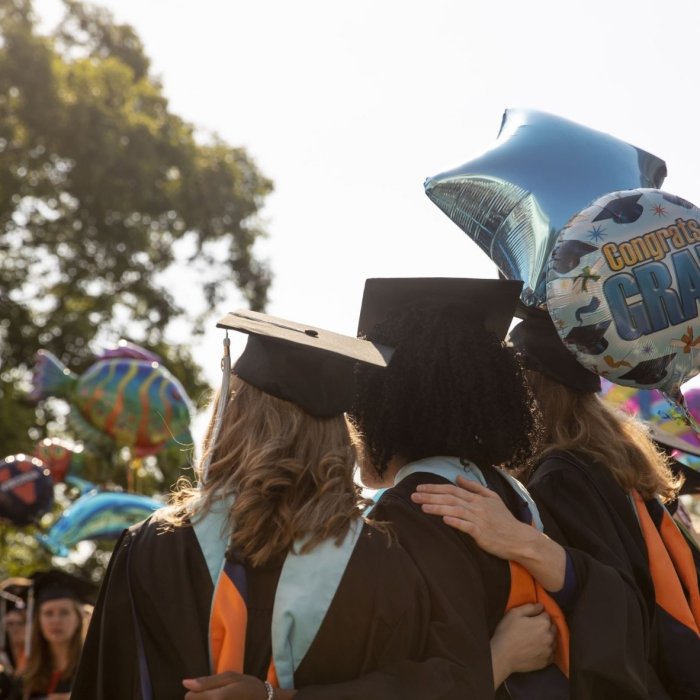 Three people wearing graduation caps and gowns with their arms around each other holding balloons