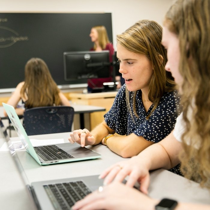 Two students sit in a classroom chatting and looking at laptops