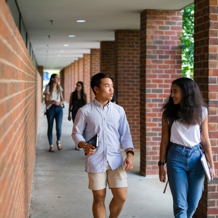 Two students chat while walking down a covered walkway next to a brick building