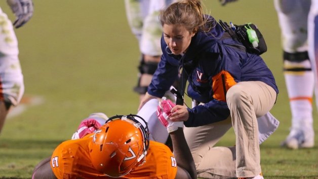 A female athletic trainer kneels over a football player in an orange jersey and helmet, who is lying on the field, and offers him water.