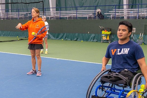 A woman wearing an orange sweatshirt stands on a tennis court holding a racket and talking. To her left, a man in a wheelchair also holds a tennis racket and looks over the net.