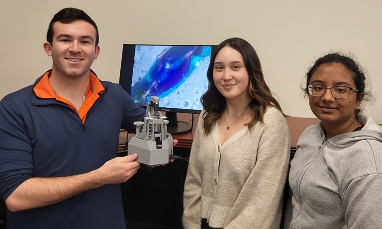 Three students stand next to each other, all smiling at the camera. The one on the left is holding a 3D printed microscope.
