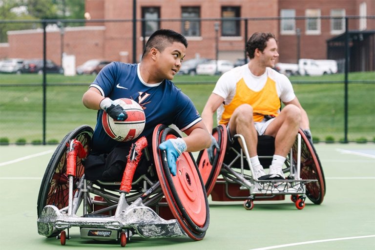 Students in sport wheelchairs play wheelchair rugby on a green outdoor tennis court