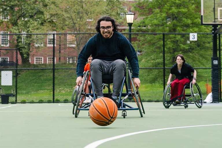 A student in a sport wheelchair speeds toward a basketball on an outdoor court