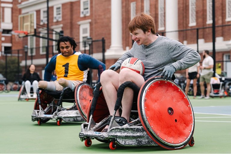 Two students in sport wheelchairs play wheelchair rugby on a green outdoor tennis court
