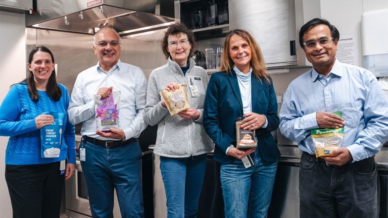 Five researchers stand in a lab kitchen, each holding a packaged food item and smiling at the camera.