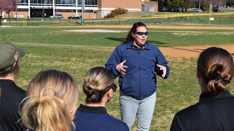 A woman stands on a baseball field speaking to several people gathered around her