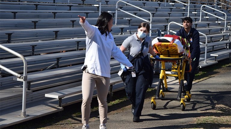 A person directs EMS personnel who are rolling a stretcher past some bleachers