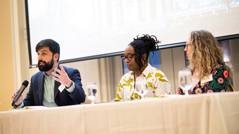 Three panelists sit behind a table, one of them speaking into a microphone.