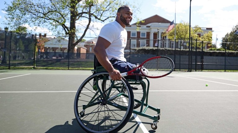 Douglas sits in a wheelchair on a tennis court with a tennis racket in his hand.