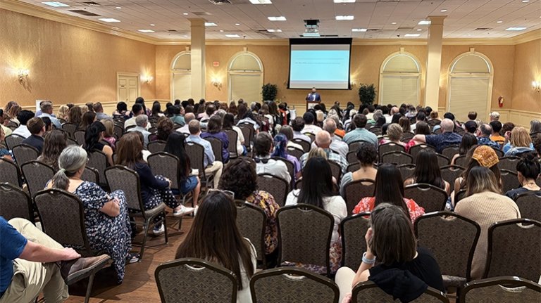 Many people sit inside at an event listening to a person speaking at a podium