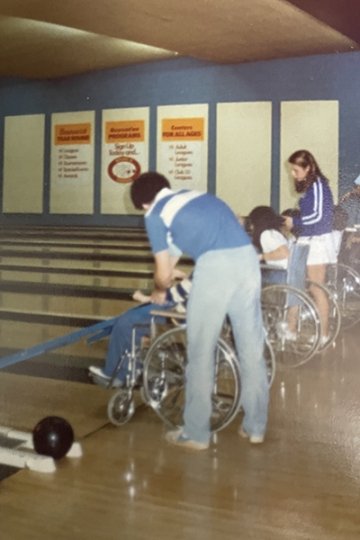 Martin Block leans over the back of a wheelchair helping the individual sitting in it hold a bowling ball.