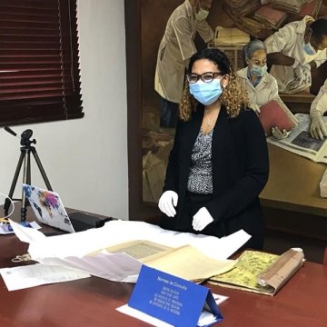 Alexa Rodriguez, wearing a mask and gloves, standing at a table with old documents and papers.