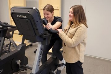 One woman uses an exercise bike while another woman stands next to her